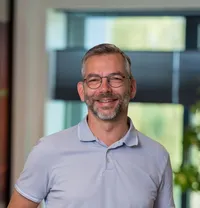 Marco Voorwinden Portret van een glimlachende man van middelbare leeftijd met kort grijs haar, baard en bril, gekleed in een lichtblauwe polo op het Twyzer kantoor met grote ramen en groen op de achtergrond.
