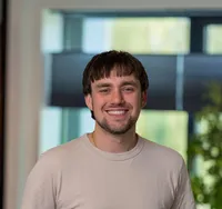 Wout den Buurman Portret van een jonge man met bruin haar, snor en sikje, gekleed in een beige T-shirt, glimlachend in een licht modern kantoor van Twyzer.