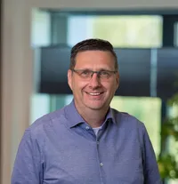 Don van Meel Portret van een man van middelbare leeftijd met kort donker haar, bril en een blauw overhemd, glimlachend in een modern licht kantoor van Twyzer.
