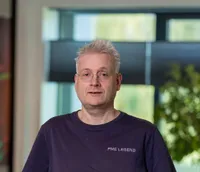 Serge Veugelers Portret van een man van middelbare leeftijd met kort grijs haar, bril en een donkerblauw T-shirt, staand neutraal kijkend op het Twyzer kantoor met ramen op de achtergrond.