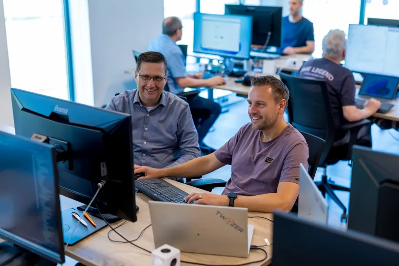 Grip op retail begint bij data die je kunt vertrouwen Twee lachende collega's aan een bureau met meerdere schermen