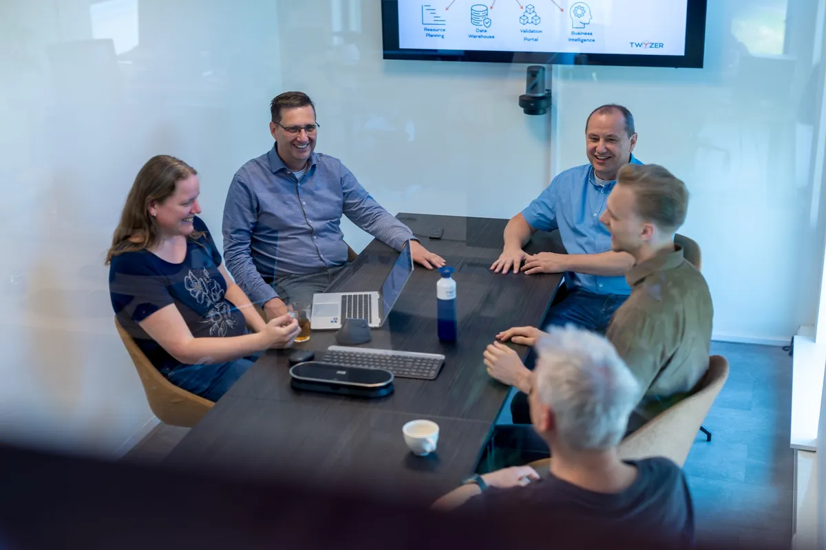 Vijf collega's zitten lachend rond een vergadertafel in een vergaderzaal met een Twyzer-presentatie op het scherm, gezien door een glazen wand.