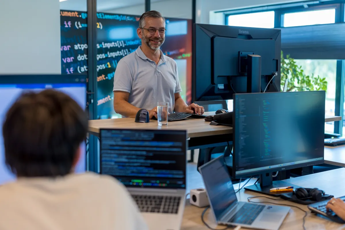 Glimlachende medewerker aan sta-bureau Een man met bril en baard staat glimlachend achter een verstelbaar bureau met meerdere schermen waarop programmeercode te zien is; op de voorgrond zit een collega aan een werkplek.