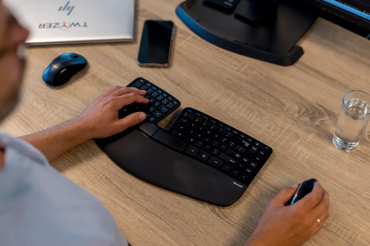 Ergonomisch toetsenbord op bureau Over-de-schouder zicht op een medewerker die werkt met een Microsoft Sculpt ergonomisch toetsenbord en muis aan een houten bureau, met een laptop met Twyzer-logo op de achtergrond.